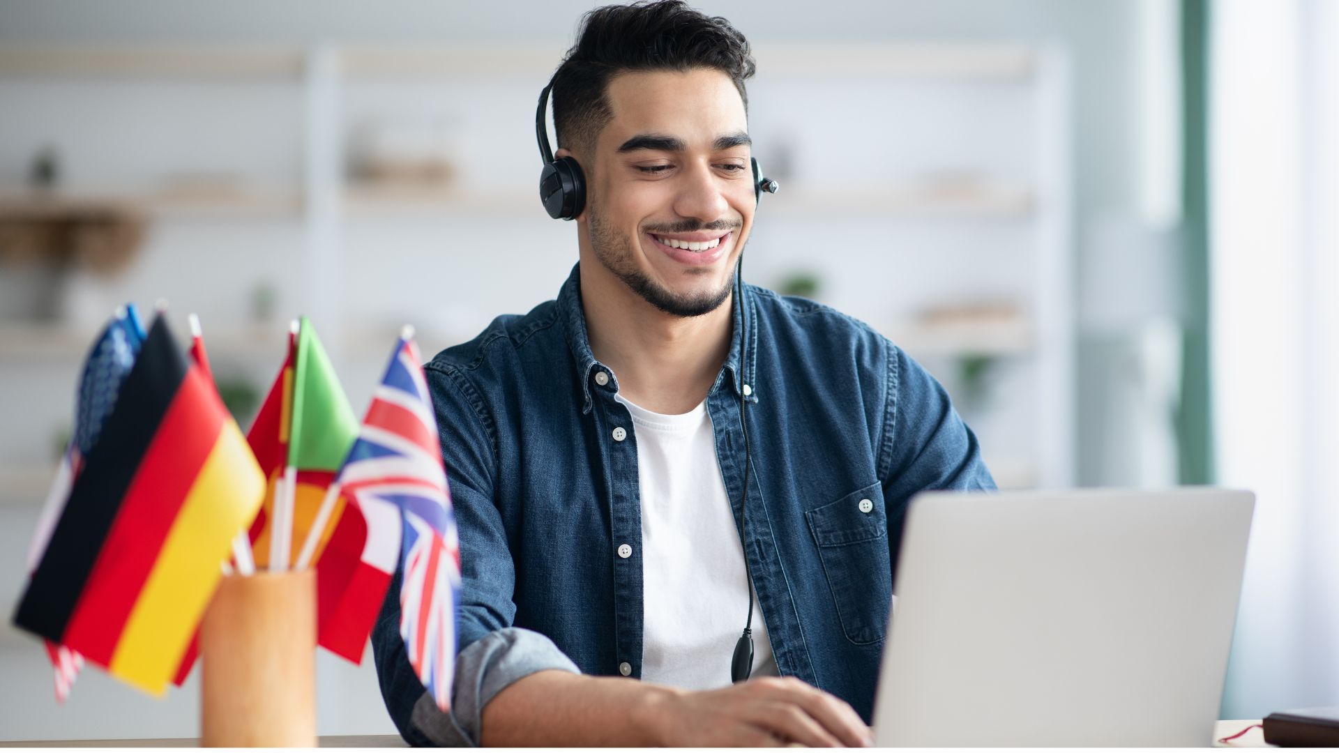 Glimlachende medewerker met headset aan laptop naast vlaggetjes van verschillende talen