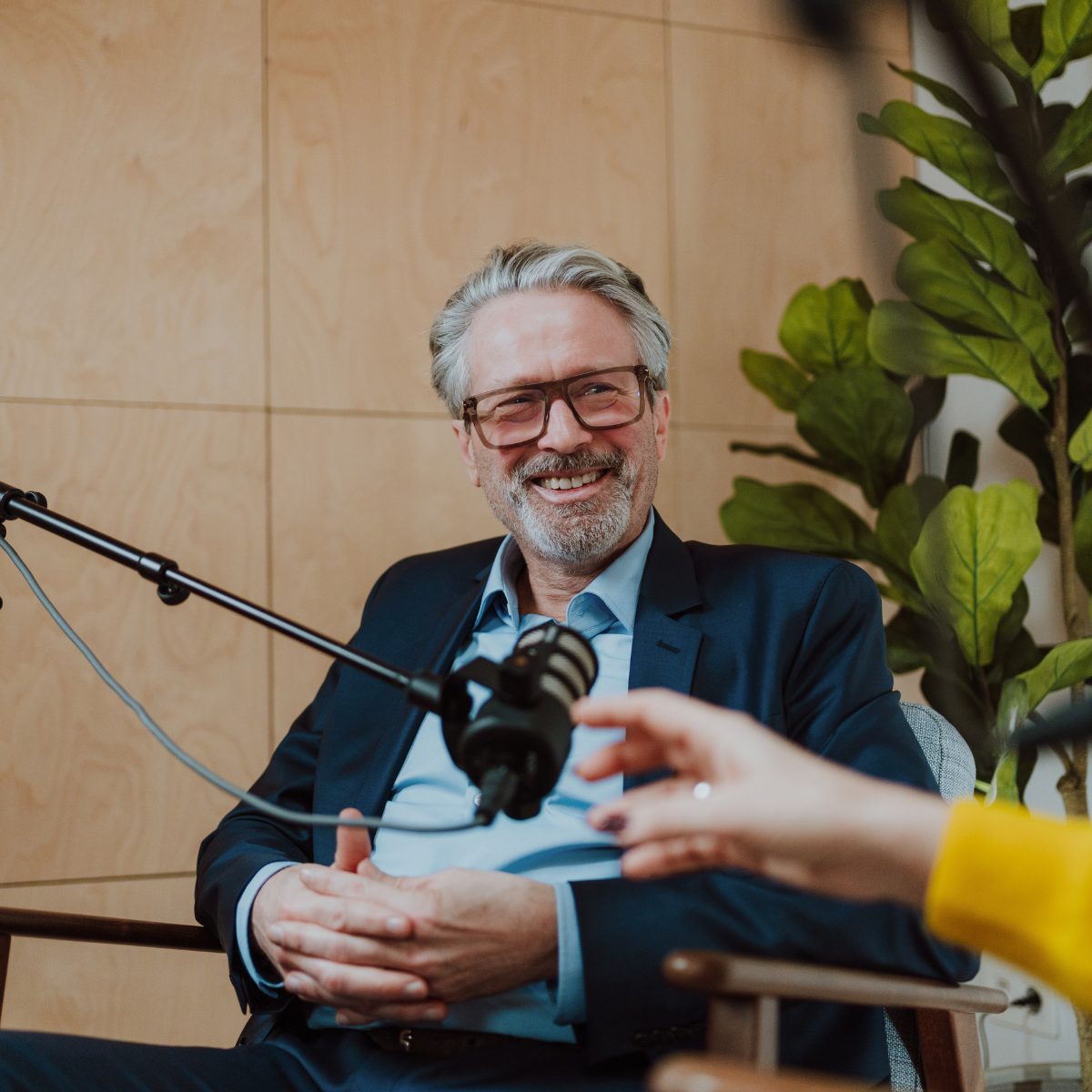 Lachende podcastgast in blauw jasje aan de microfoon tijdens een klankbordsessie