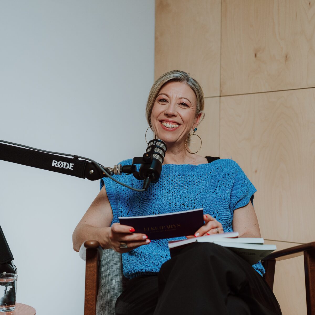 Lachende Elke Van Parys in blauwe trui aan de RØDE-microfoon in de podcaststudio