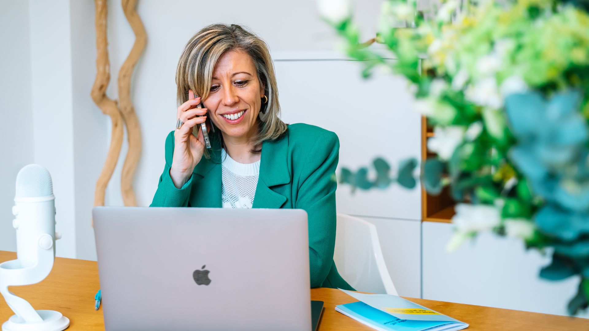 Vrouw in groene blazer aan de telefoon achter laptop in een lichte kantooromgeving
