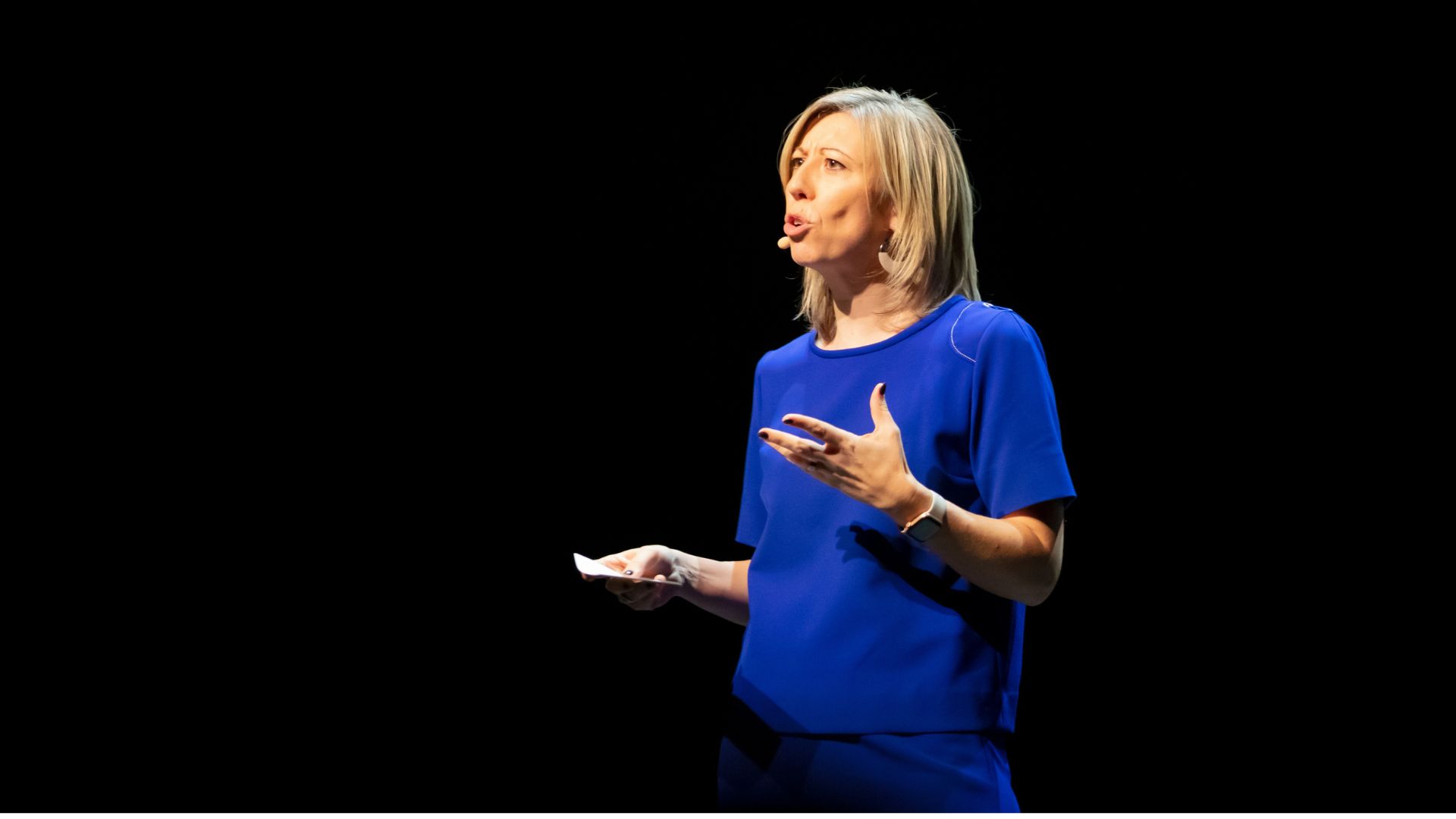 Elke Van Parys spreekt op een podium in een blauwe outfit tegen een donkere achtergrond