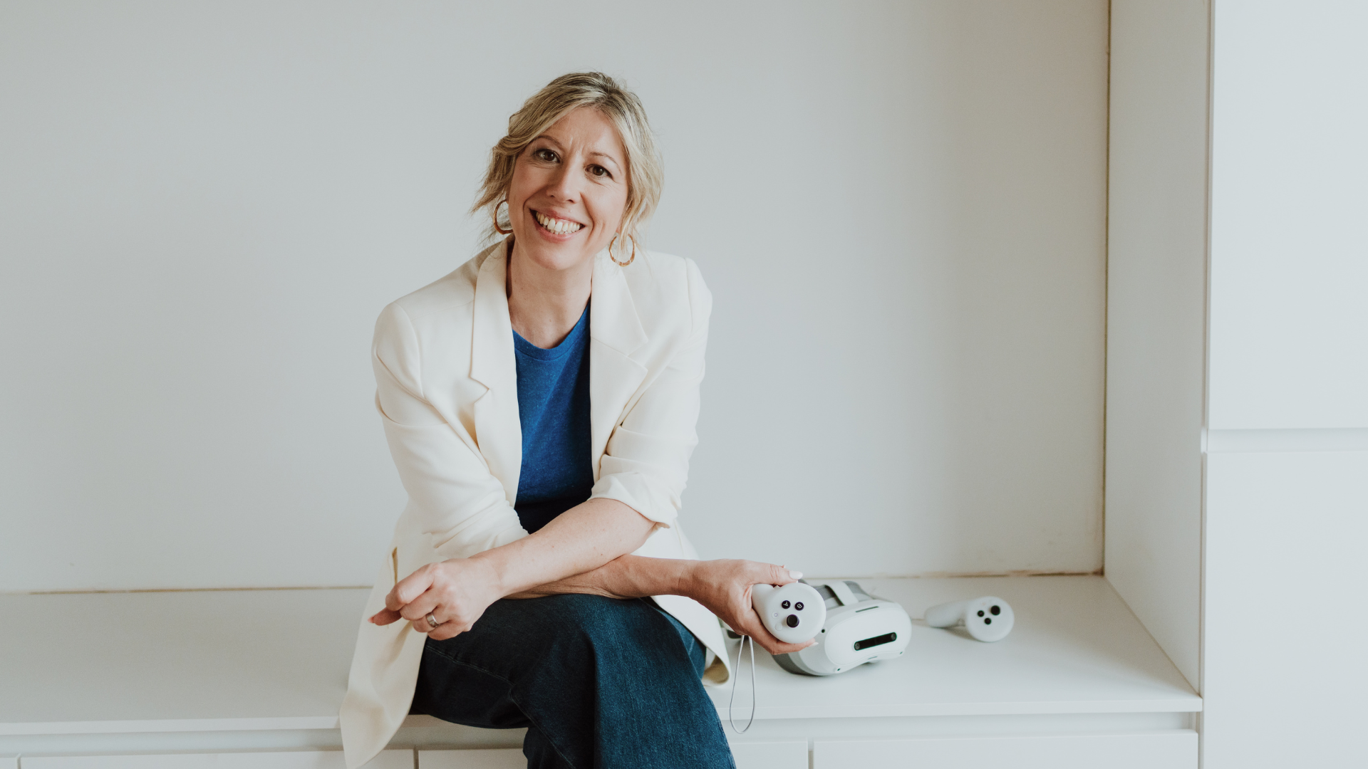 Elke Van Parys in een crèmekleurige blazer en blauwe top, zittend met een VR-headset in de hand in een lichte ruimte